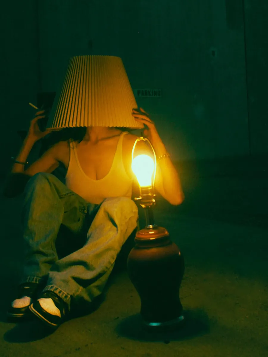 Figure wearing a lampshade beside a lit lantern in a green room